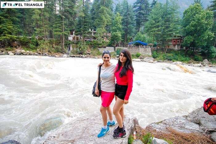 Young women travelers pose on the banks of a river flowing through Kasol