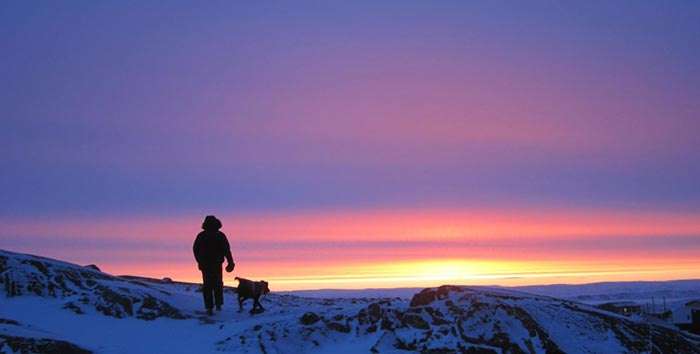 A man walks his dog in the snow in Iqaluit in evening while the sun still shines