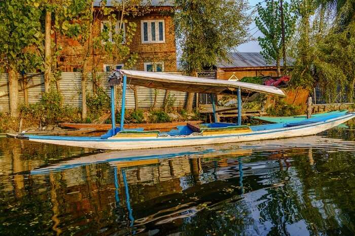 Old rugged wooden boat floated by an old house on a shore of Dal Lake during sunset