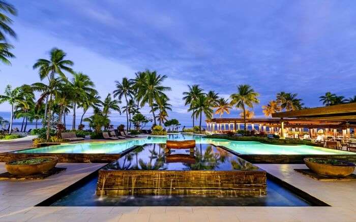 A lavish pool surrounded by palm trees overlooking a beach 
