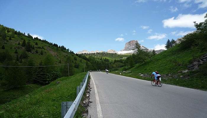 A person cycling on an empty path with a beautiful view of green valleys and Dolomite peaks in the background.