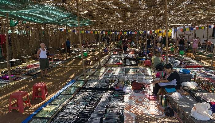 Busy scene at Anjuna Flea Market with colourful handicraft, garment, and jewellery stalls, interspersed with shoppers.