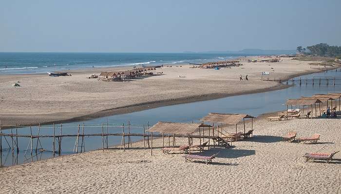 A panoramic view of Mandrem Beach showing white sands, blue waters, bamboo bridges crossing over a small creek, and palm trees at the back near the Paliem Sweet Water Lake.