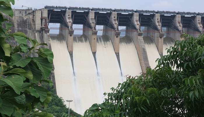 Picturesque view of Sardar Sarovar Dam with the gigantic Statue of Unity at the back, against a clear blue sky, amidst lush greenery and the serene Narmada River. 